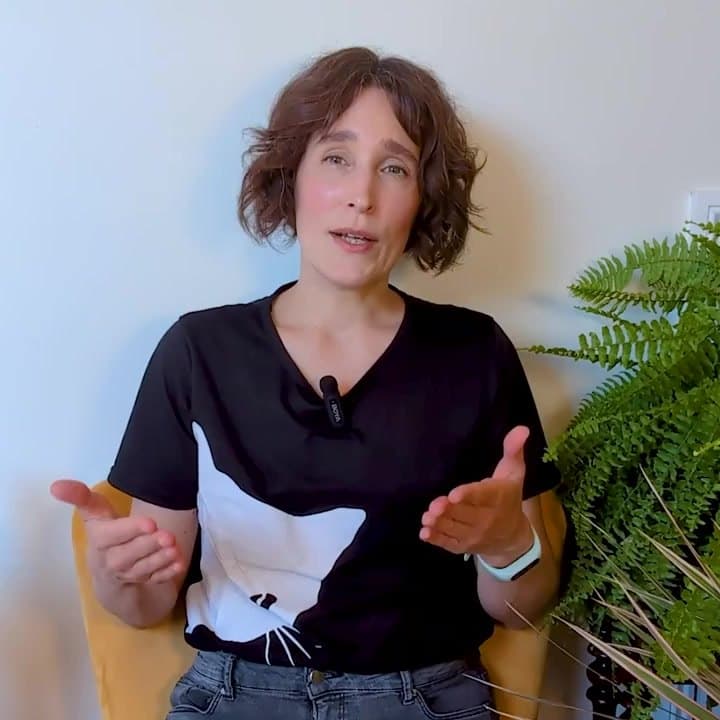 Kristin Loeer, a woman with short curly brown hair in a black T-shirt, sitting in a yellow chair beside a fern, talking with her hands.