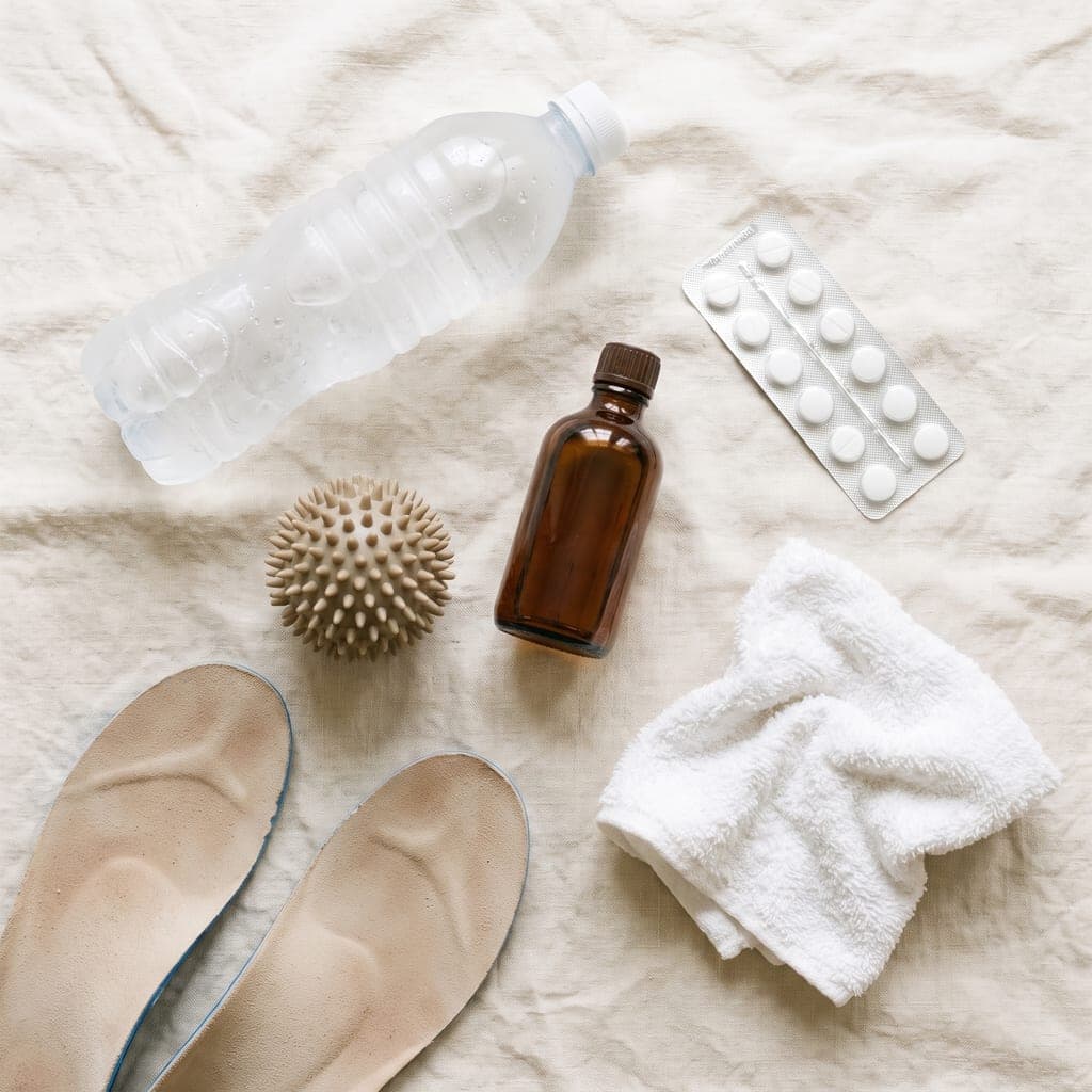 A frozen water bottle, a spiky massage ball, a small bottle of massage oil, a blister pack of tablets, a pair of arch-support insoles and a scrunched hand towel, laid out on pale linen.