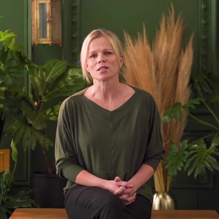 Theresa Moodie, a woman with short fair hair, in an olive top, sitting against a green panelled wall with plants.