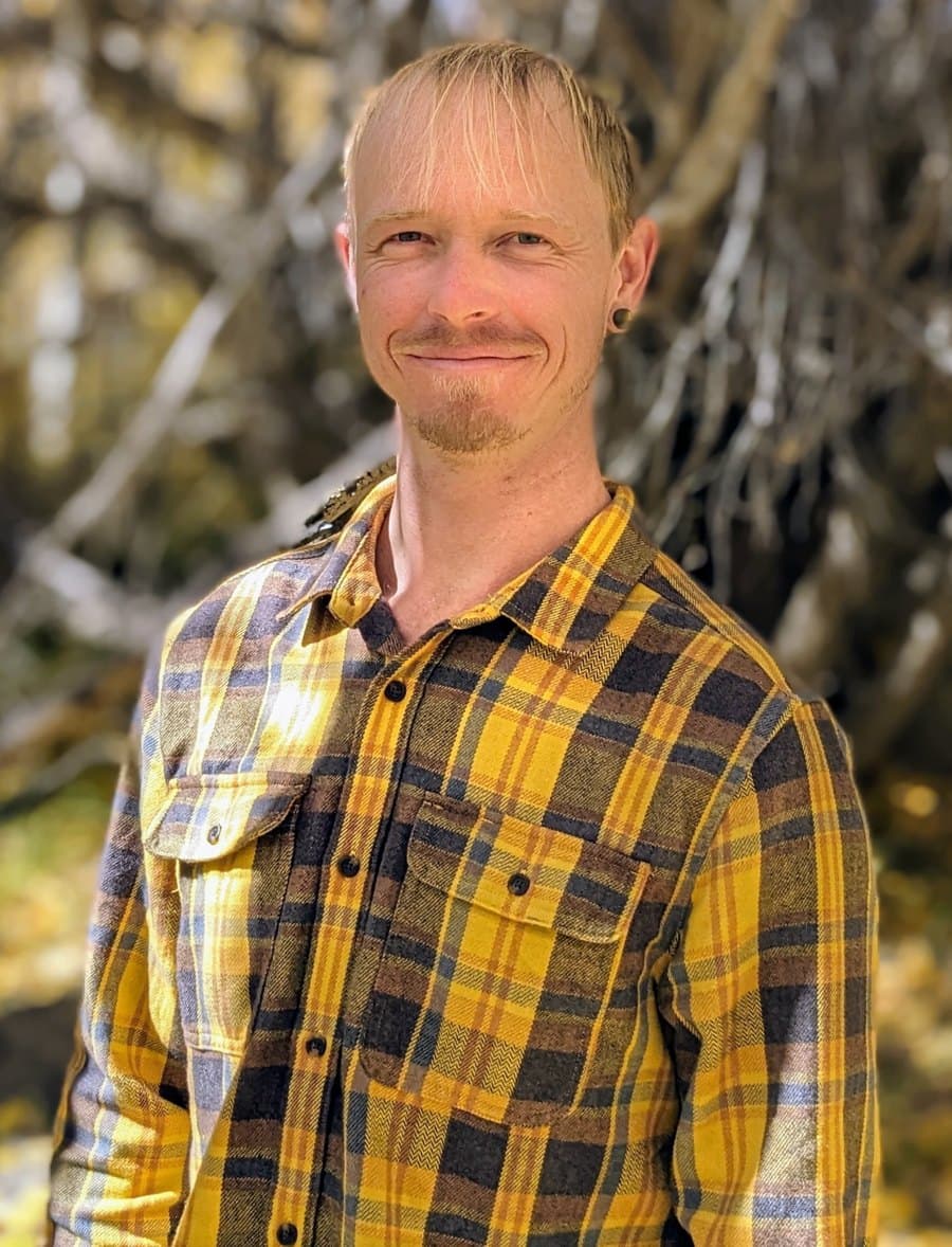 Asa Dean, a man with short fair hair and a short beard in a yellow and navy plaid shirt, smiling, standing among bare branches outdoors.